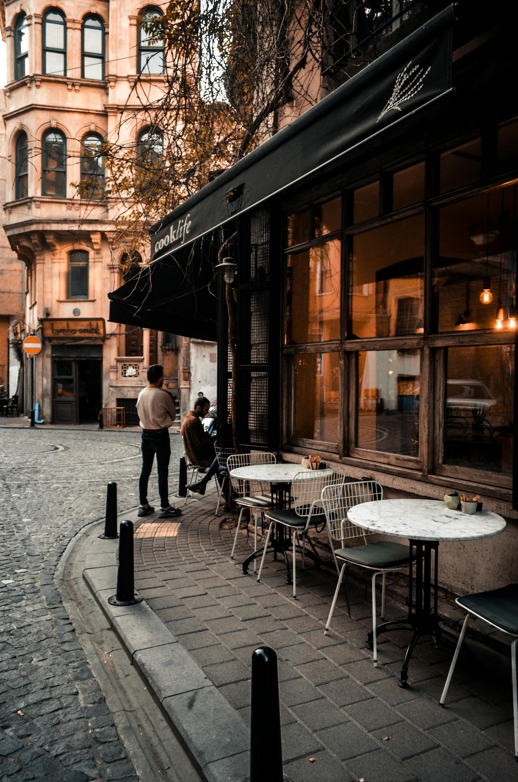 Facade of cafe with large windows and tables on sidewalk along road and faceless people resting in city
